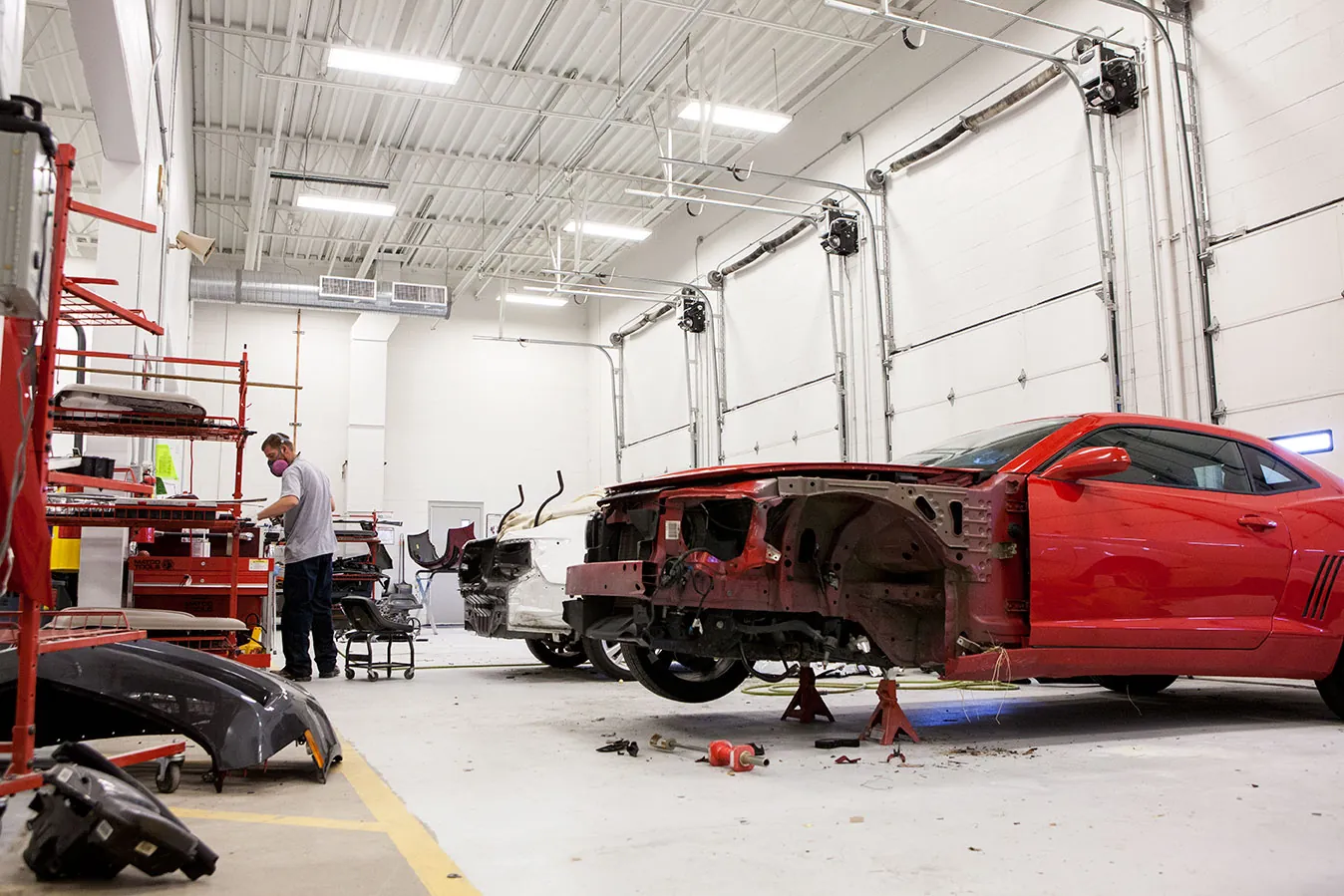 A red Camaro mid-repair on the shop floor with a technician at work