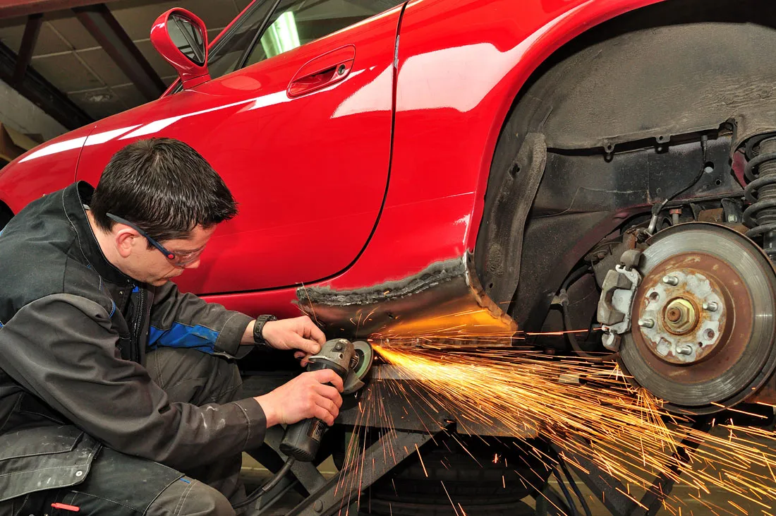 A technician grinding a rocker panel on a red sports car, sparks flying