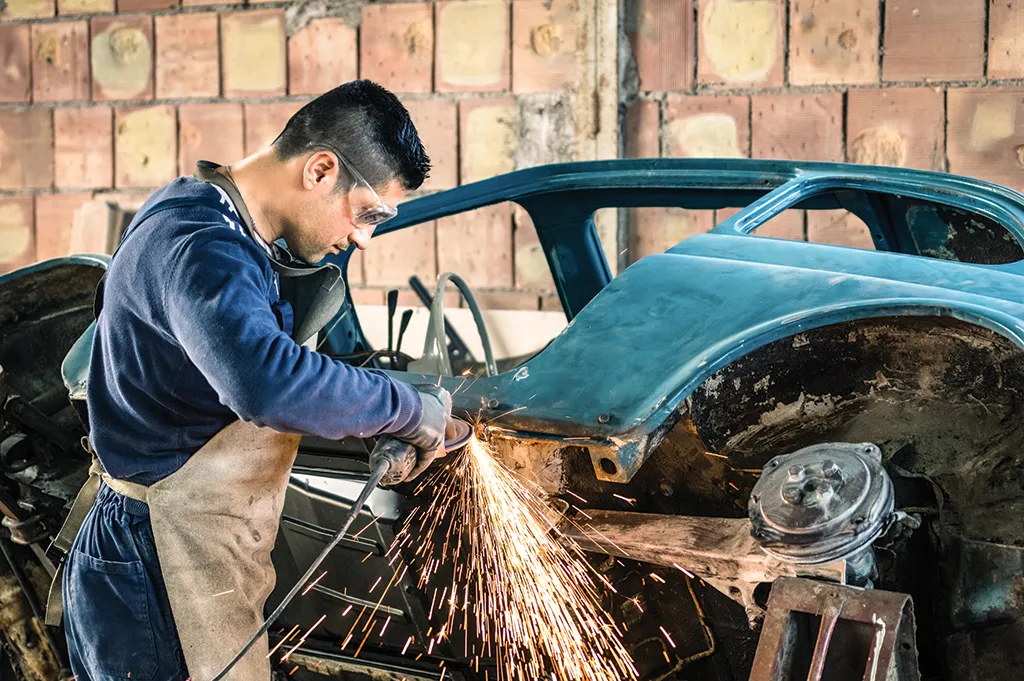 A technician grinding bodywork on a vintage blue car against a brick wall